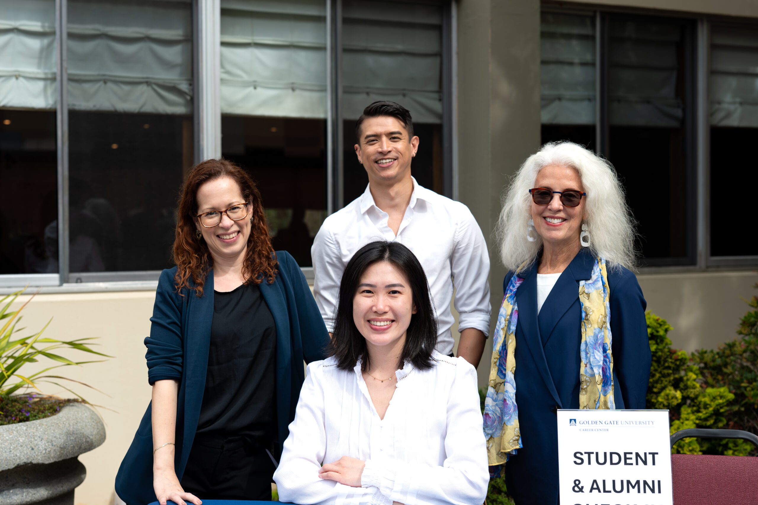 Four smiling people stand and sit in front of a building with large windows and light gray awnings, with green shrubs and a stone planter at the edges. A woman with curly auburn hair and glasses, wearing a navy cardigan over black, leans on the table at left. A man in a white open-collared shirt stands behind them at center top. A woman with long white hair, sunglasses and a blue floral scarf, wearing a navy blazer, stands at right. In front, a woman with shoulder-length dark hair in a white blouse sits with her arms crossed. A white sign in the lower right reads “Golden Gate University Career Center” and “Student & Alumni Outreach” (partly cut off).