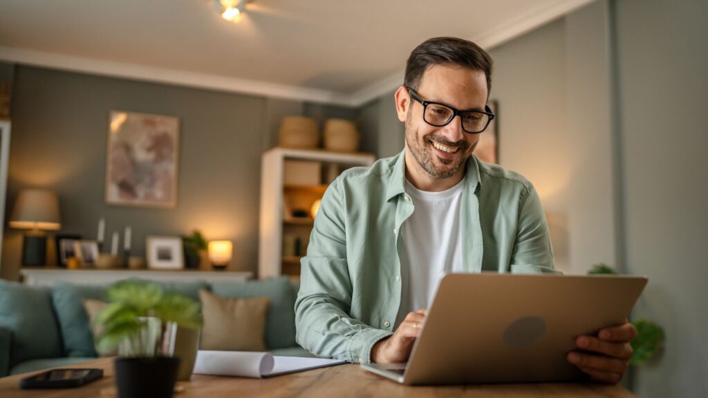 Smiling man sitting at a table in his home and looking at a laptop.
