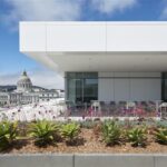 Rooftop terrace with landscaped planters, outdoor seating, and views of San Francisco City Hall in the background. The terrace is framed by a modern white building façade under a partly cloudy sky.