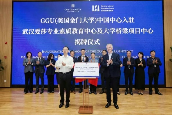 Two men in business attire stand next to a document, with a row of professionals lined up behind them. They are clapping in celebration. A blue sign with white lettering, naming both partners, serves as a backdrop.