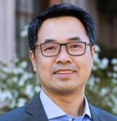 A middle-aged Asian man with black hair and black framed glasses smiles. He wears a light blue button up under a navy blazer, in front of a garden.