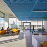 Light-filled study lounge with large windows, yellow curved seating, and city views. Students work individually and meet in small groups around tables and lounge areas.