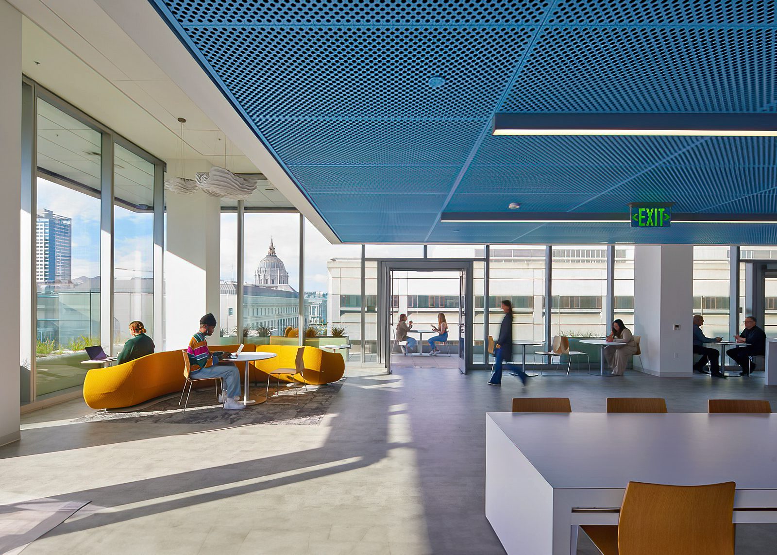 Light-filled study lounge with large windows, yellow curved seating, and city views. Students work individually and meet in small groups around tables and lounge areas.