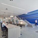 Contemporary academic commons featuring a long communal worktable, pendant lighting, and a striking blue striped feature wall beside an open staircase. A student works on a laptop while others move through the space.