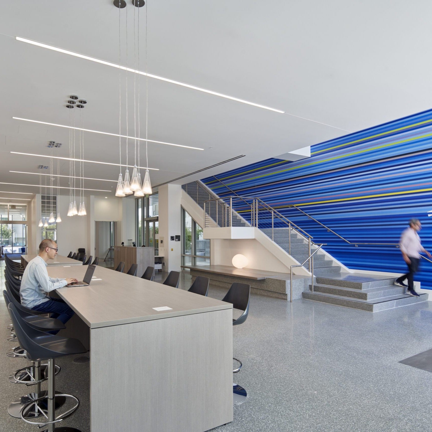 Contemporary academic commons featuring a long communal worktable, pendant lighting, and a striking blue striped feature wall beside an open staircase. A student works on a laptop while others move through the space.