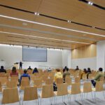 Modern lecture hall with rows of light wood chairs facing a large projection screen. A presenter stands at the front while students are seated throughout the room beneath a wood-paneled ceiling with integrated lighting.