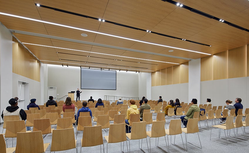 Modern lecture hall with rows of light wood chairs facing a large projection screen. A presenter stands at the front while students are seated throughout the room beneath a wood-paneled ceiling with integrated lighting.