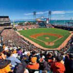 An aerial view of a packed baseball stadium during a daytime game, with crowds of fans dressed in orange and black filling the stands. The green diamond and brown infield are visible at center, with San Francisco Bay, sailboats, and distant hills visible beyond the outfield under a bright blue sky.