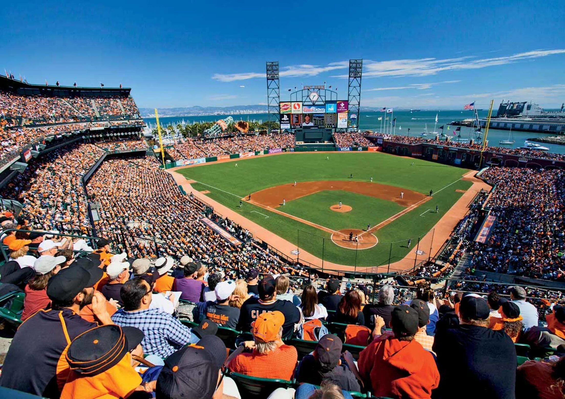 An aerial view of a packed baseball stadium during a daytime game, with crowds of fans dressed in orange and black filling the stands. The green diamond and brown infield are visible at center, with San Francisco Bay, sailboats, and distant hills visible beyond the outfield under a bright blue sky.