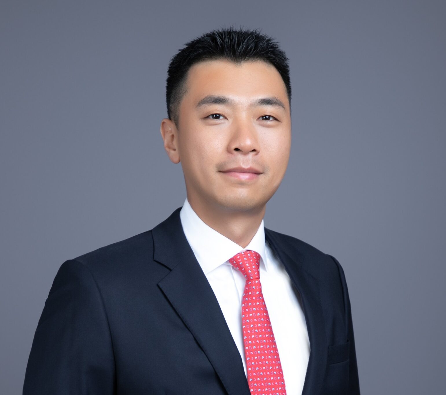 Professional headshot of Benson Lu against a neutral gray background. He is wearing a dark navy suit jacket, white dress shirt, and red patterned tie, with hands in his pockets. He is looking directly at the camera with a composed expression.