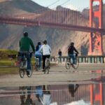 A group of cyclists wearing helmets ride along a waterfront path toward the Golden Gate Bridge, with the bridge's red towers and the Marin Headlands reflected in a puddle on the path in the foreground.