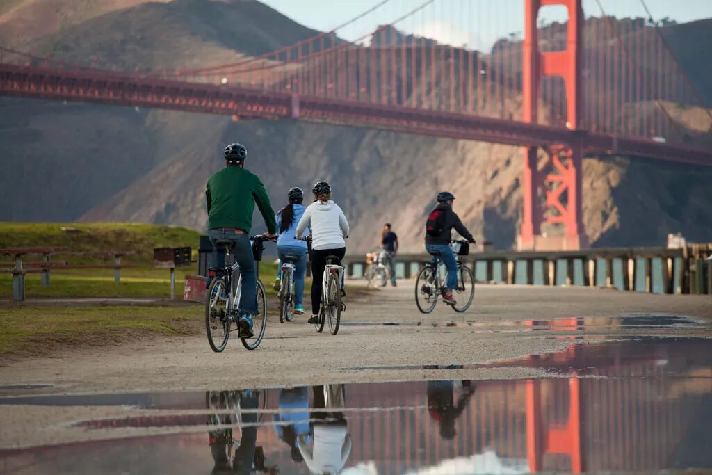 A group of cyclists wearing helmets ride along a waterfront path toward the Golden Gate Bridge, with the bridge's red towers and the Marin Headlands reflected in a puddle on the path in the foreground.