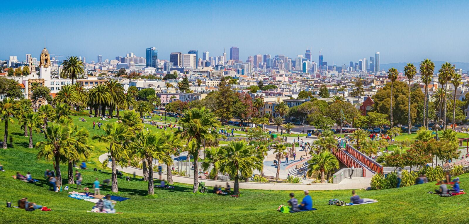 A sunny afternoon at Dolores Park in San Francisco, with people relaxing and picnicking on the grassy hillside among palm trees, a playground and pathways below, and the city's downtown skyline stretching across the horizon under a clear blue sky.