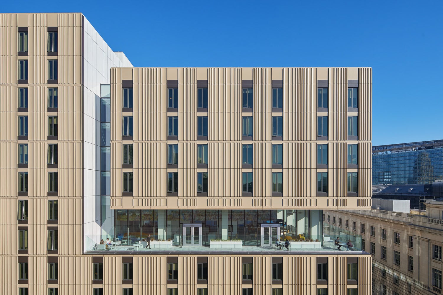 Close-up exterior view of GGU's Downtown Campus at The Academe at 198 in San Francisco. The contemporary high-rise features a light-colored metal façade with vertical architectural fins, large rectangular windows, and a recessed outdoor terrace with glass railings. Several people are visible using the terrace space. The building is framed against a clear blue sky, with neighboring downtown buildings visible in the background.