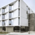 Modern academic building with white architectural panels, expansive windows, and concrete detailing at a street corner.