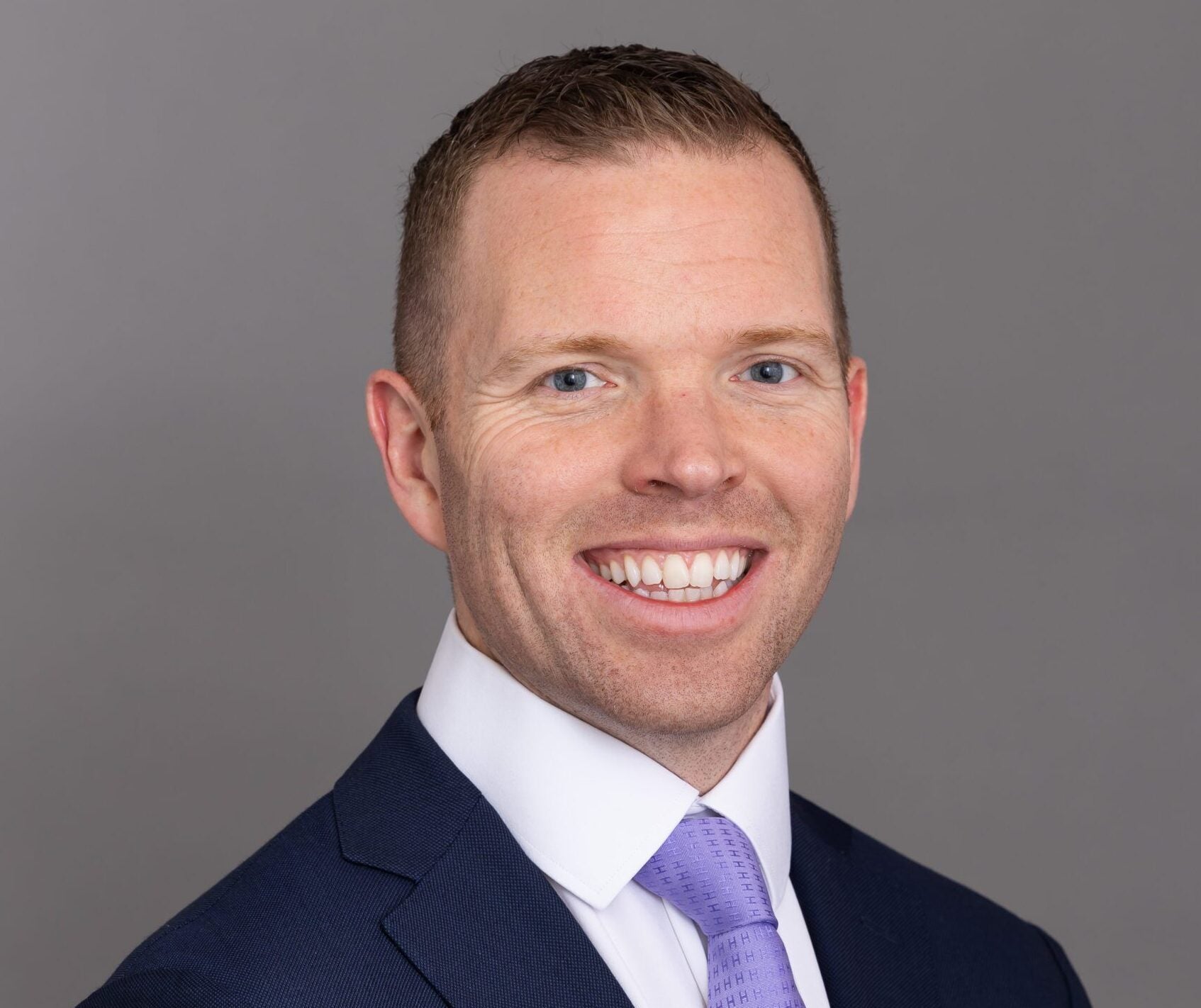 Professional headshot of a smiling man in a navy blue suit, white dress shirt, and light purple tie, photographed against a neutral gray background.