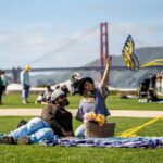 Two people relax on a blanket in a grassy park on a sunny day, with a woman smiling and holding up a butterfly-shaped kite with yellow ribbon tails, a picnic basket of flowers beside them, and the Golden Gate Bridge visible in the background.