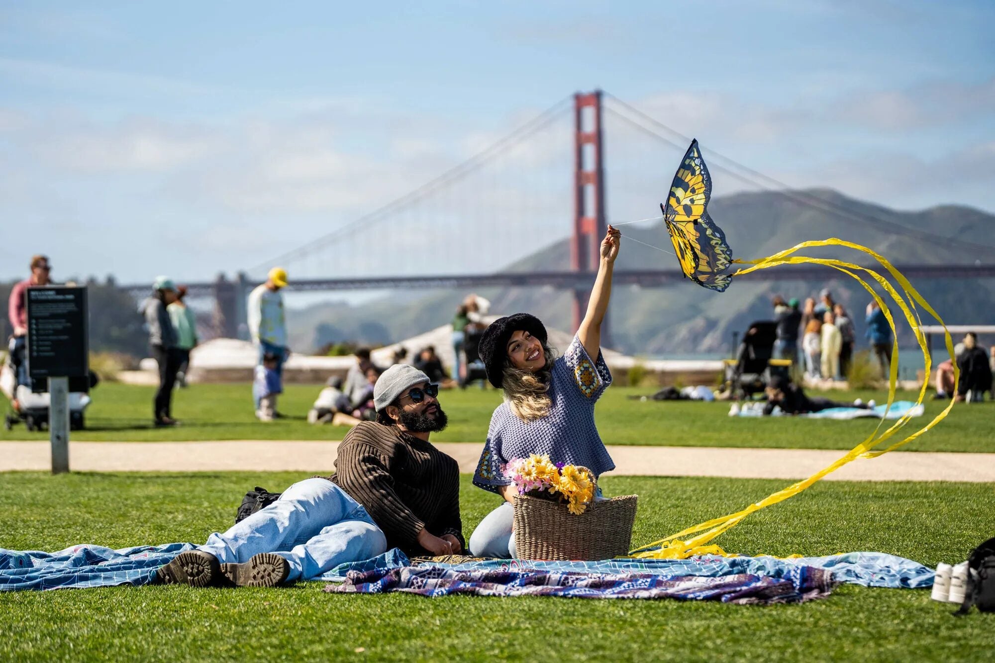 Two people relax on a blanket in a grassy park on a sunny day, with a woman smiling and holding up a butterfly-shaped kite with yellow ribbon tails, a picnic basket of flowers beside them, and the Golden Gate Bridge visible in the background.
