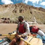 A man in a red life jacket and camo cap rows a gear-laden raft down a muddy river through a rugged desert canyon, holding yellow oars and looking out across the water beneath a blue sky with scattered clouds.