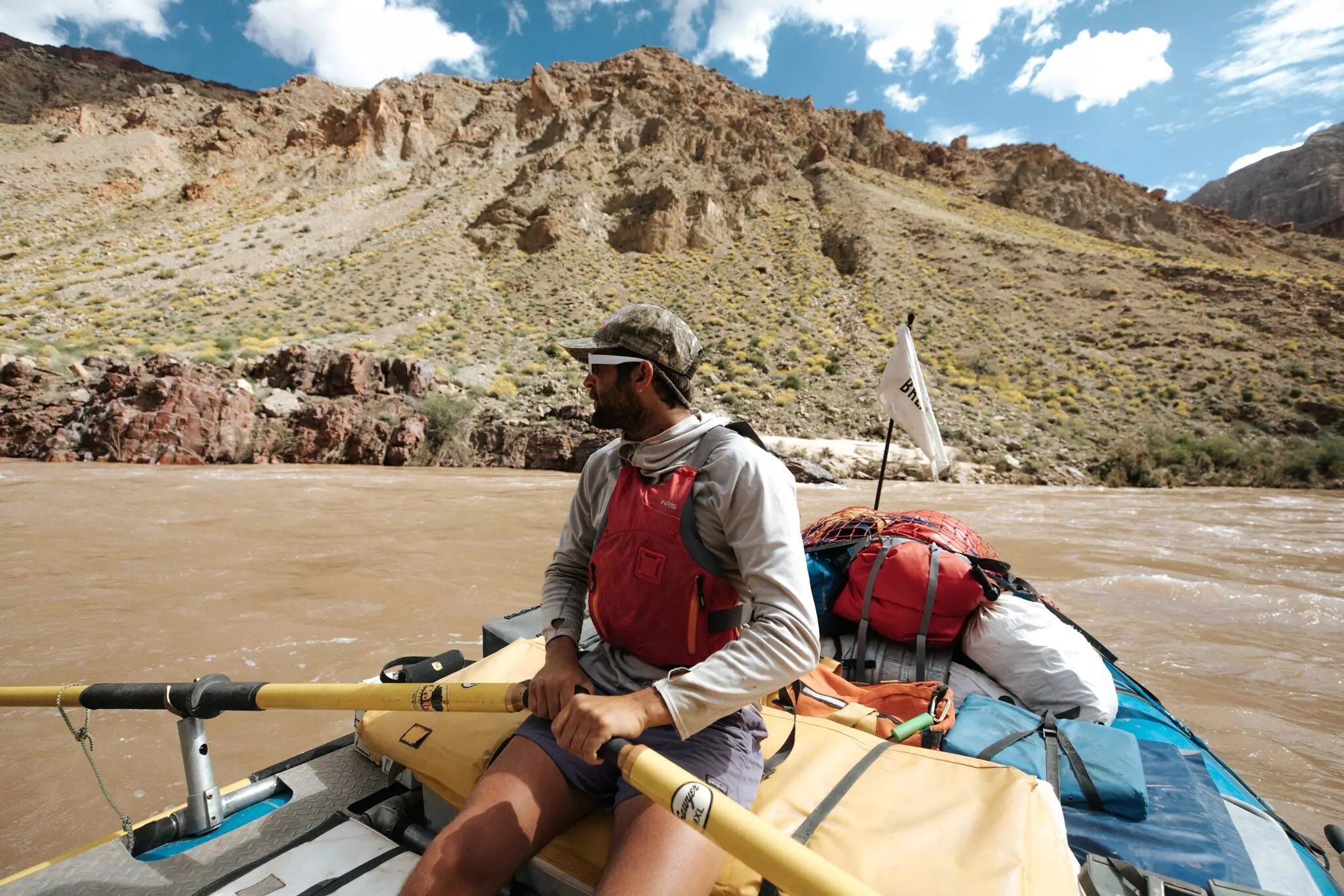 A man in a red life jacket and camo cap rows a gear-laden raft down a muddy river through a rugged desert canyon, holding yellow oars and looking out across the water beneath a blue sky with scattered clouds.