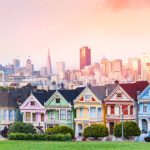A row of colorfully painted Victorian houses known as the Painted Ladies at Alamo Square, featuring vivid hues of green, red, yellow, and blue, with the San Francisco skyline including the Transamerica Pyramid visible behind them beneath a warm pink and peach sunset sky.