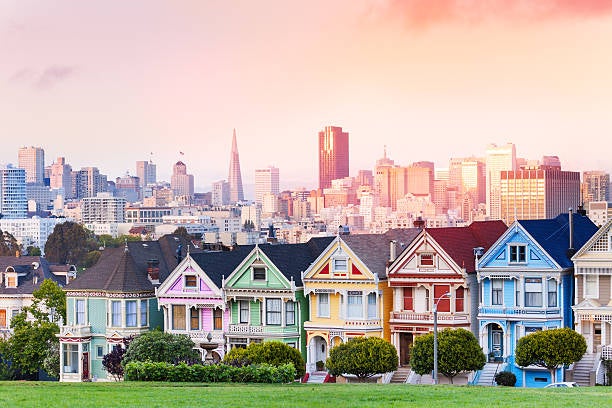 A row of colorfully painted Victorian houses known as the Painted Ladies at Alamo Square, featuring vivid hues of green, red, yellow, and blue, with the San Francisco skyline including the Transamerica Pyramid visible behind them beneath a warm pink and peach sunset sky.