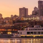 Aerial view of Ghirardelli Square and the San Francisco waterfront at sunset, with the illuminated Ghirardelli sign glowing orange against a warm golden sky, a small red sailboat moored in the foreground, and the city's hillside neighborhoods rising in the background.