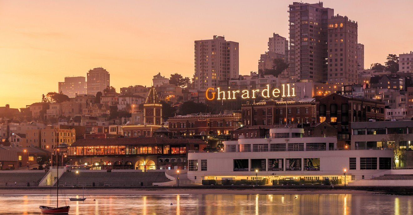 Aerial view of Ghirardelli Square and the San Francisco waterfront at sunset, with the illuminated Ghirardelli sign glowing orange against a warm golden sky, a small red sailboat moored in the foreground, and the city's hillside neighborhoods rising in the background.