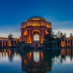 The Palace of Fine Arts in San Francisco illuminated in warm amber light at twilight, its grand rotunda and colonnades reflected in the still lagoon in front, with a deep blue sky and silhouetted trees framing the scene.