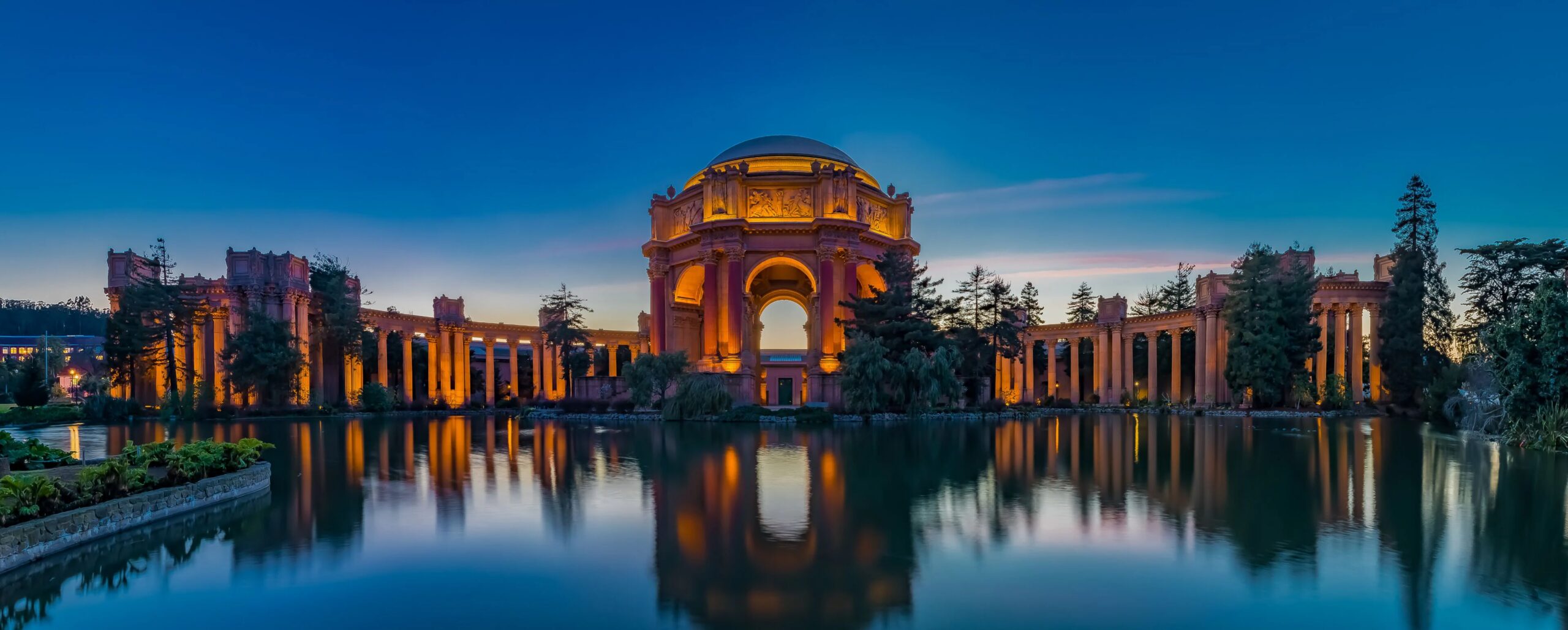 The Palace of Fine Arts in San Francisco illuminated in warm amber light at twilight, its grand rotunda and colonnades reflected in the still lagoon in front, with a deep blue sky and silhouetted trees framing the scene.