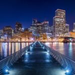 A long pier extending toward the brightly lit San Francisco skyline at night, with colorful reflections of city lights shimmering across the calm bay waters on both sides, and a deep blue sky overhead.