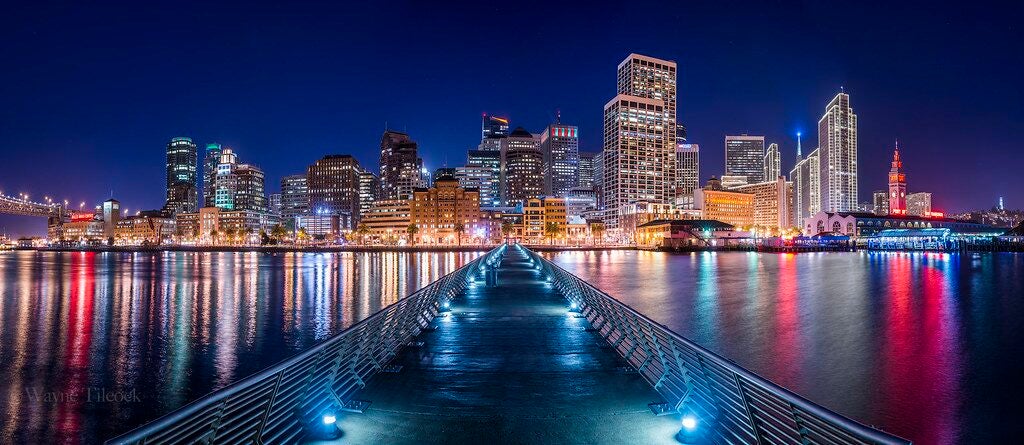 A long pier extending toward the brightly lit San Francisco skyline at night, with colorful reflections of city lights shimmering across the calm bay waters on both sides, and a deep blue sky overhead.
