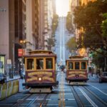 Two classic San Francisco cable cars traveling along California Street in opposite directions, with the road rising steeply uphill toward a golden sunrise and tall city buildings lining both sides of the street.