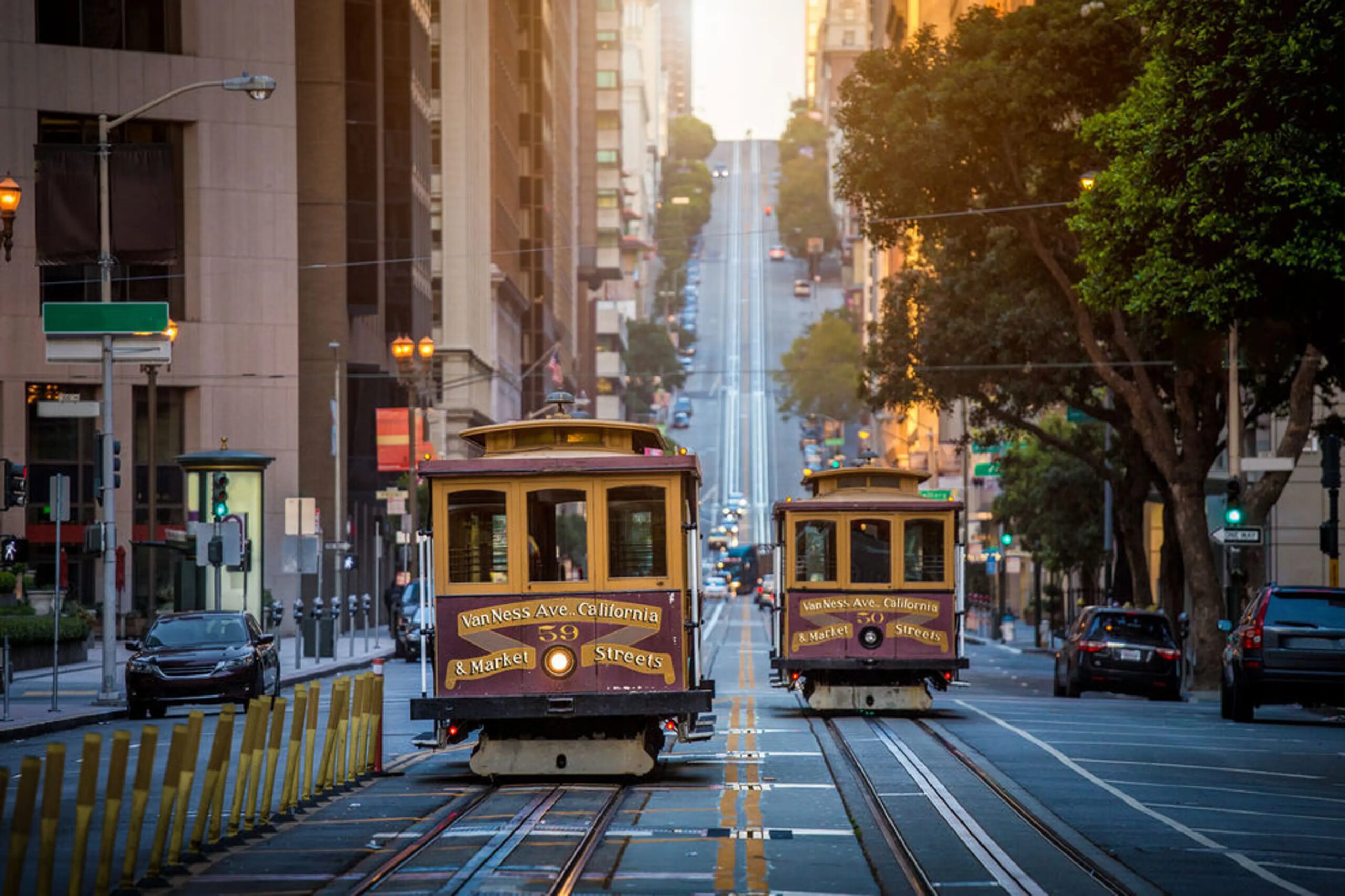 Two classic San Francisco cable cars traveling along California Street in opposite directions, with the road rising steeply uphill toward a golden sunrise and tall city buildings lining both sides of the street.