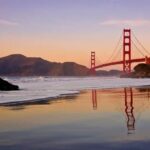 The Golden Gate Bridge reflected in the wet sand of a beach at sunset, its red towers glowing against a warm peach and lavender sky, with gentle waves washing ashore and the Marin Headlands visible in the distance.