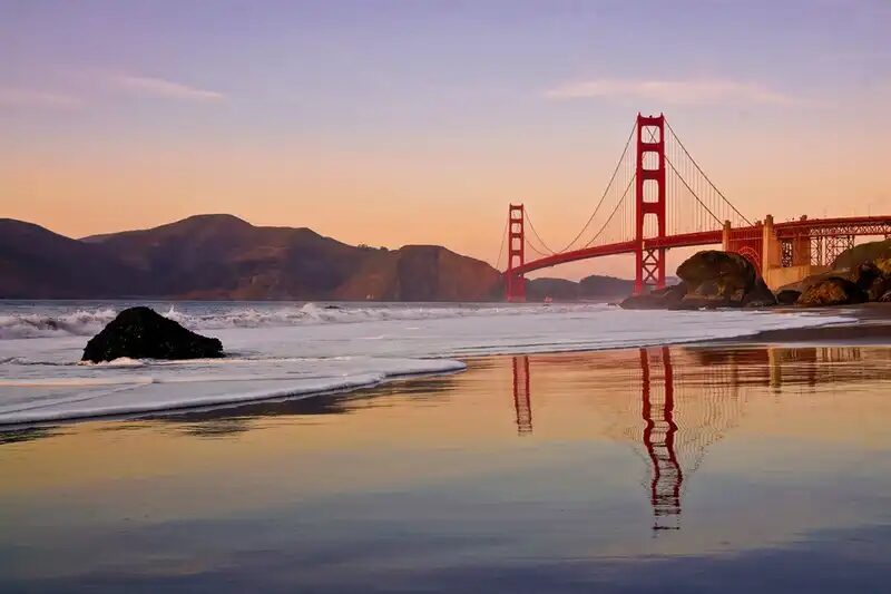 The Golden Gate Bridge reflected in the wet sand of a beach at sunset, its red towers glowing against a warm peach and lavender sky, with gentle waves washing ashore and the Marin Headlands visible in the distance.