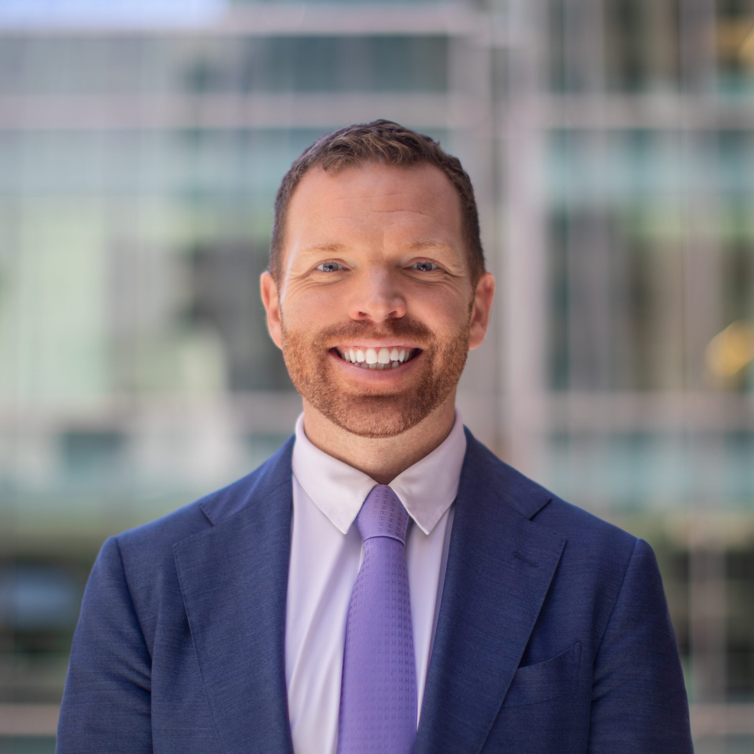 Professional headshot of Taylor Jantz, smiling in a navy suit and purple tie, with a blurred glass office building in the background.