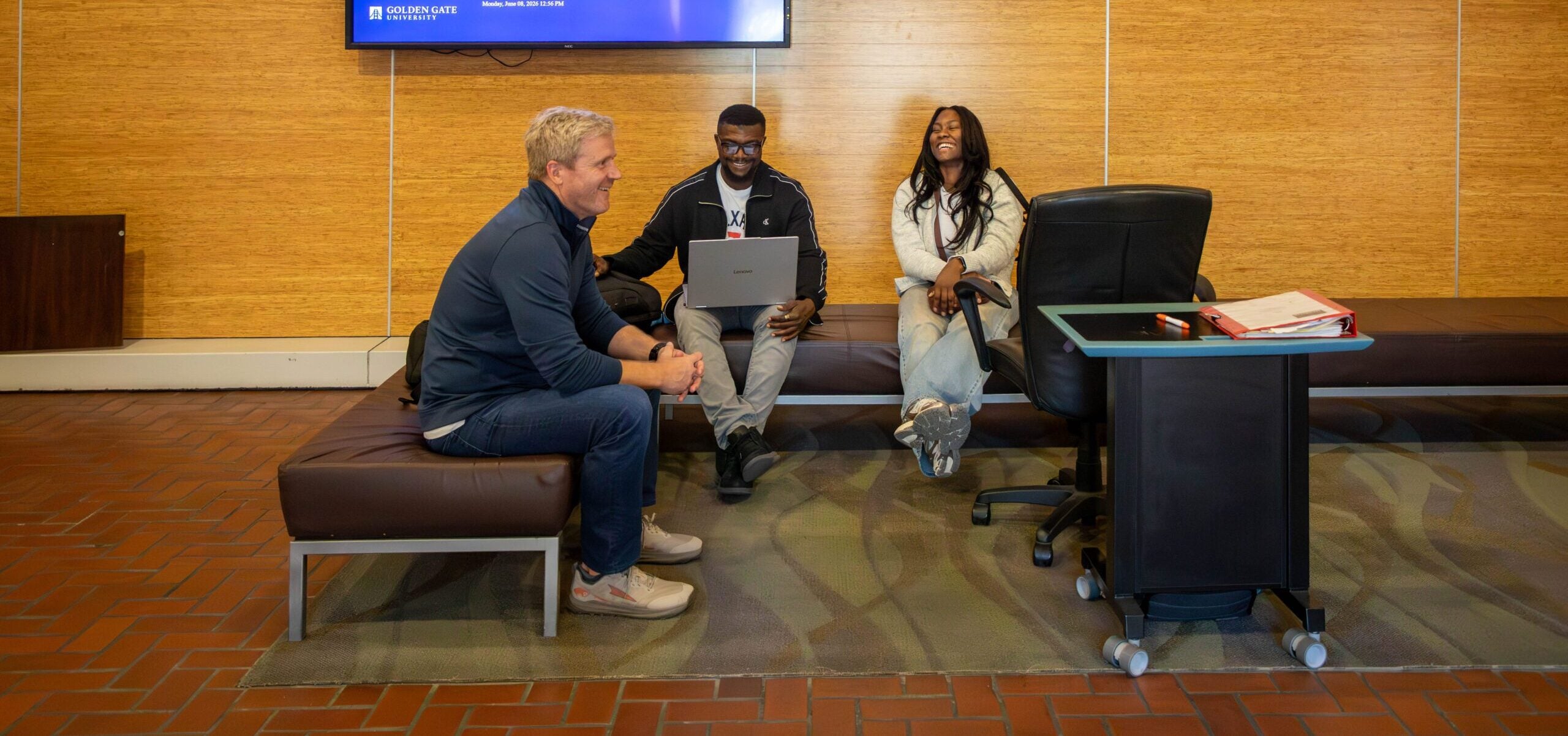 Three people laughing together on a bench seating area, one with a laptop, under a wall-mounted screen displaying a 