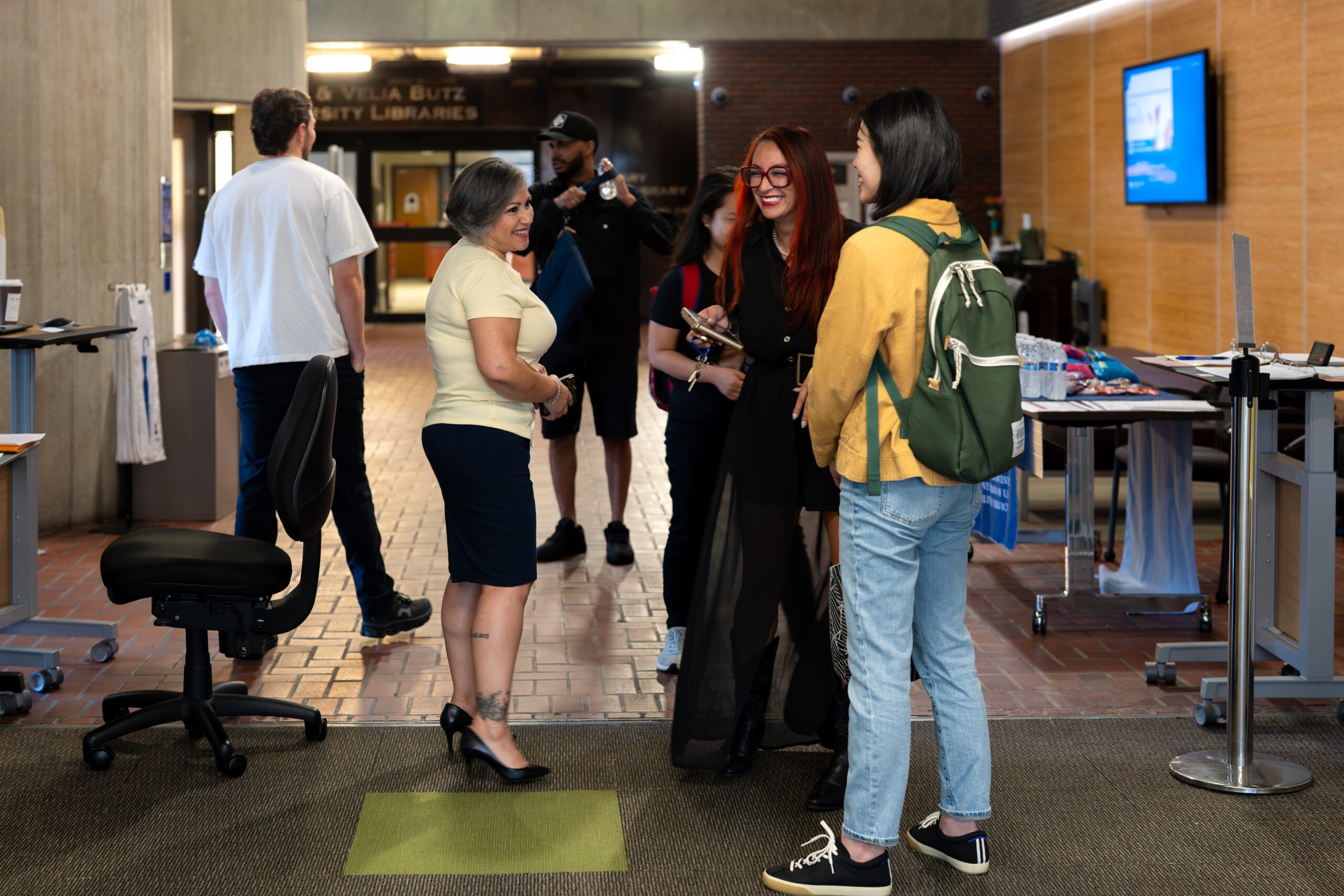 A small group of students and staff chatting and smiling near a table with water bottles and snacks, in a lobby area outside the university library entrance.