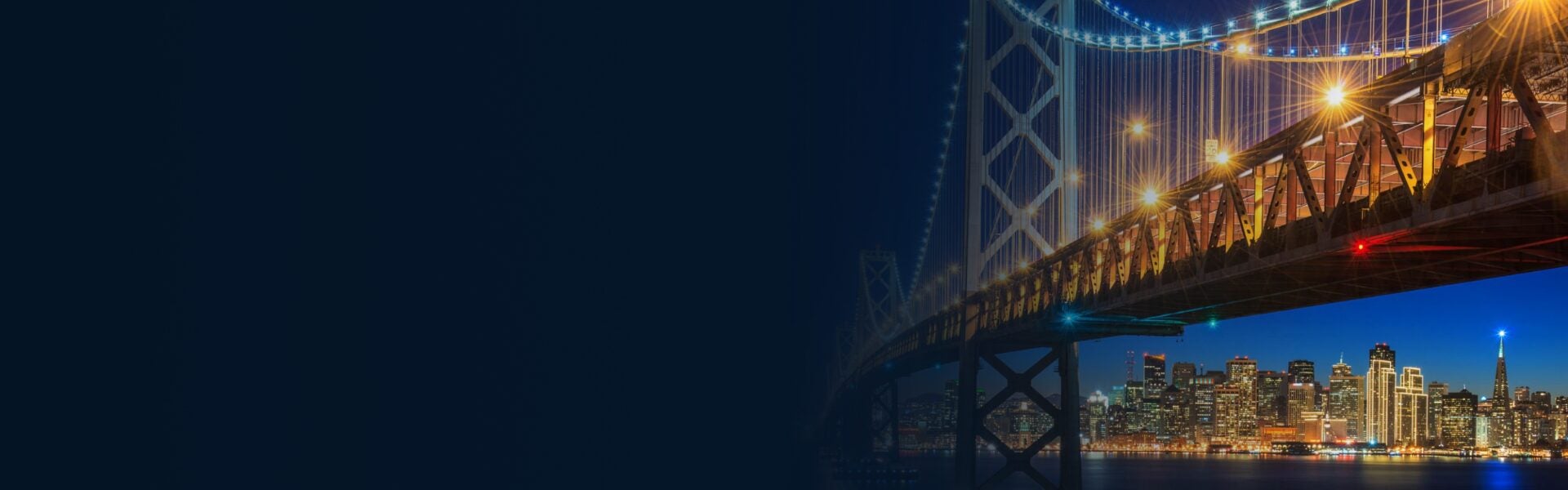 Night view of the San Francisco Bay Bridge's illuminated steel truss span arching across the frame, with the glowing downtown skyline visible beneath it against a deep blue twilight sky.