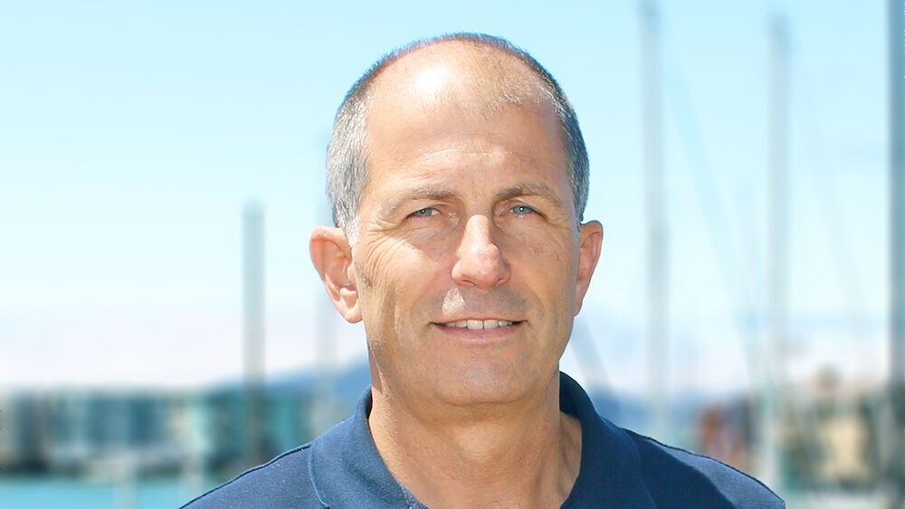 an with grey hair wearing a dark blue polo shirt, photographed outdoors with a waterfront and blurred urban skyline in the background.