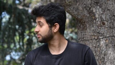 Man with dark hair and beard wearing a black long-sleeved shirt with a white X crossed by a red rose graphic, standing in front of a blurred tree trunk and foliage.