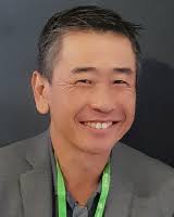 Smiling man with short gray-streaked dark hair, wearing a gray blazer over a dark shirt with a green lanyard around his neck, photographed against a dark background.