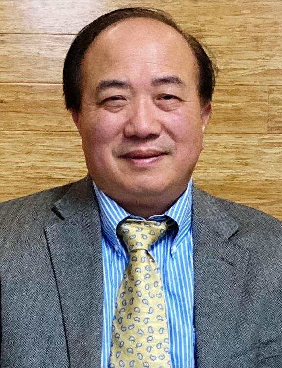 Professional headshot of a man wearing a gray blazer, blue striped dress shirt, and yellow patterned tie against a light wood paneling background.