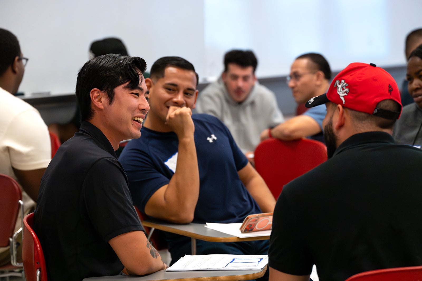 A classroom of men seated at desks with red chairs. In the foreground, a smiling man in a black polo laughs while looking toward a man in a red cap and black shirt seen from behind. Beside them, a man in a navy athletic shirt rests his chin on his hand and smiles. Papers and an orange phone case sit on the desk, and other participants are blurred in the background.
