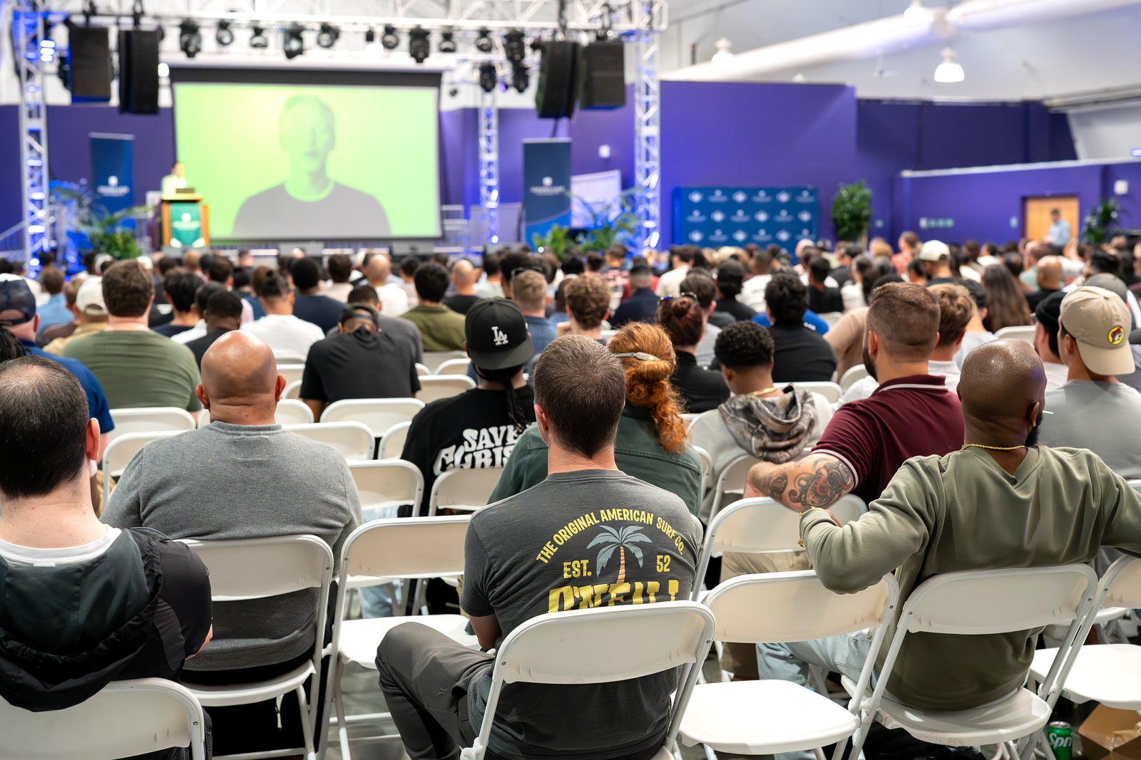 A large audience seated in white folding chairs, viewed from behind, faces a stage. A large screen shows a blurred, green-tinted image of a speaker in a gray shirt, and a speaker stands at a green podium at the far left. The purple walls are decorated with blue banners and a step-and-repeat backdrop.