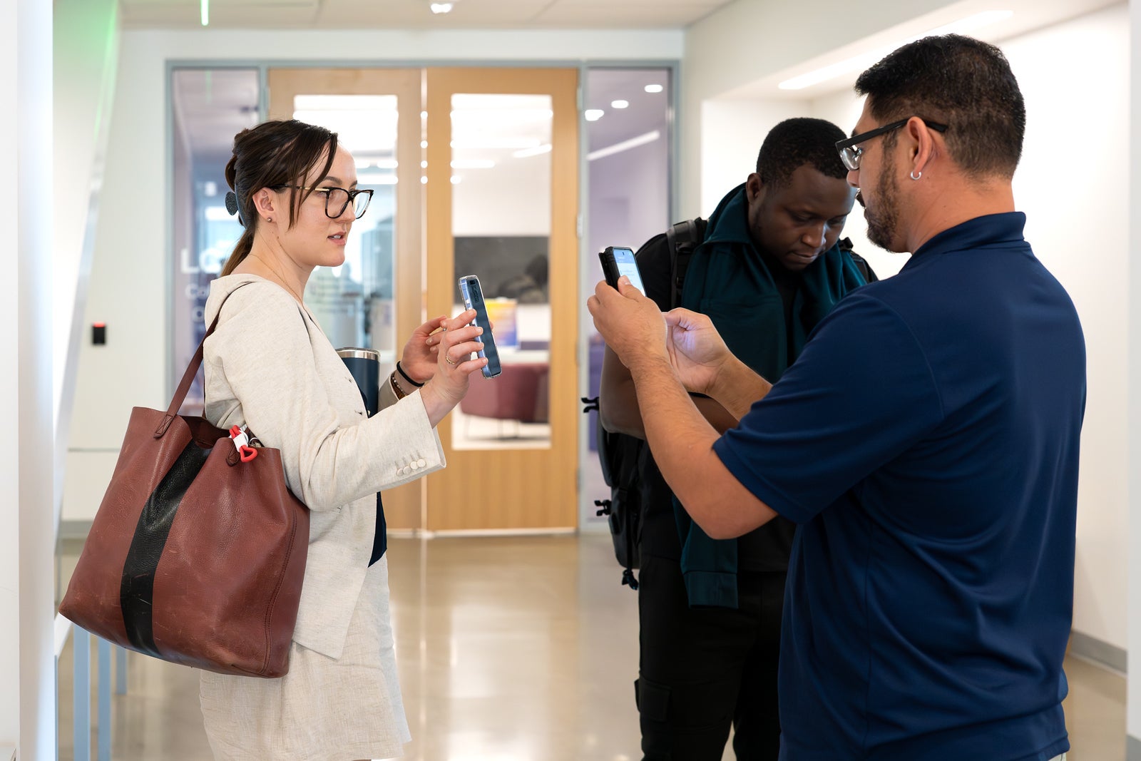 A woman in a beige blazer and brown leather tote holds up a smartphone while speaking to a man in a navy polo, who also holds a phone. A second man in a dark jacket looks down at his phone. They stand in a bright hallway near wooden doors and glass walls.