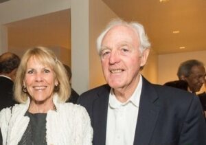 John and Barbara Davids smile at an indoor Golden Gate University event. Barbara, on the left, has shoulder-length blonde hair and wears a white textured cardigan over a dark top. John, on the right, has white hair and wears a navy blazer over a white open-collared shirt. Other guests are visible in the background.